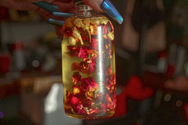 Close-up of a hand holding a glass bottle of natural facial oil surrounded by fresh herbs and flowers.