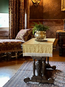An ornate room features a wooden table with intricate carvings, topped with a fringed gold tablecloth. A silver pot with lush green ferns sits on the table. In the background, a plush chaise lounge is upholstered in a rich burgundy and gold fabric, while heavy drapes match its opulent design. The walls display a textured, dark brown pattern, and a decorative wall sconce adds to the vintage feel. A patterned rug covers the wooden floor.