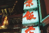Neon signs with red Chinese characters are displayed against a teal background, illuminated in a vibrant light. The signs are set near an intricately decorated building façade that features colorful architectural details.