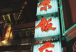 Neon signs with red Chinese characters are displayed against a teal background, illuminated in a vibrant light. The signs are set near an intricately decorated building façade that features colorful architectural details.
