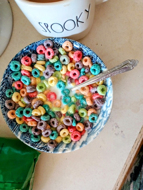A bowl filled with colorful, circular cereal pieces in a variety of colors including red, blue, green, yellow, and purple, mixed with milk. A silver spoon rests in the bowl, which features a blue and white pattern on its rim. In the background, there is a white mug with the word 'SPOOKY' printed on it. Part of a green package is visible at the bottom of the image.