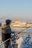 A man wearing a blue cap and a patterned sweater holds a fishing rod while standing beside a railing. Behind him, a white passenger ferry with yellow accents sails on calm water under a clear blue sky. Bags and other fishing equipment are attached to the railing.