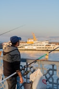 A man wearing a blue cap and a patterned sweater holds a fishing rod while standing beside a railing. Behind him, a white passenger ferry with yellow accents sails on calm water under a clear blue sky. Bags and other fishing equipment are attached to the railing.