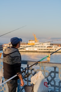 A man wearing a blue cap and a patterned sweater holds a fishing rod while standing beside a railing. Behind him, a white passenger ferry with yellow accents sails on calm water under a clear blue sky. Bags and other fishing equipment are attached to the railing.