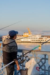A man wearing a blue cap and a patterned sweater holds a fishing rod while standing beside a railing. Behind him, a white passenger ferry with yellow accents sails on calm water under a clear blue sky. Bags and other fishing equipment are attached to the railing.