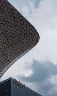 An architectural structure with a hexagonal pattern curves elegantly against a backdrop of a cloudy sky. Below, a modern building displays the logo 'Telcel' prominently.