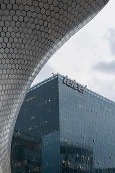 A modern building with a hexagonal patterned structure casts a unique, curved facade over a glass skyscraper with a visible brand name 'telcel' on its top corner. The sky is overcast, suggesting a cloudy day.