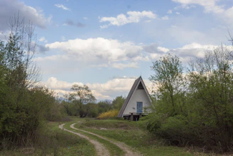A triangular A-frame house is situated in a natural setting, surrounded by lush green trees and bushes. A narrow dirt path leads to the house, winding through the grassy landscape. Above, the sky is partly cloudy, with patches of blue visible amidst the fluffy white clouds.
