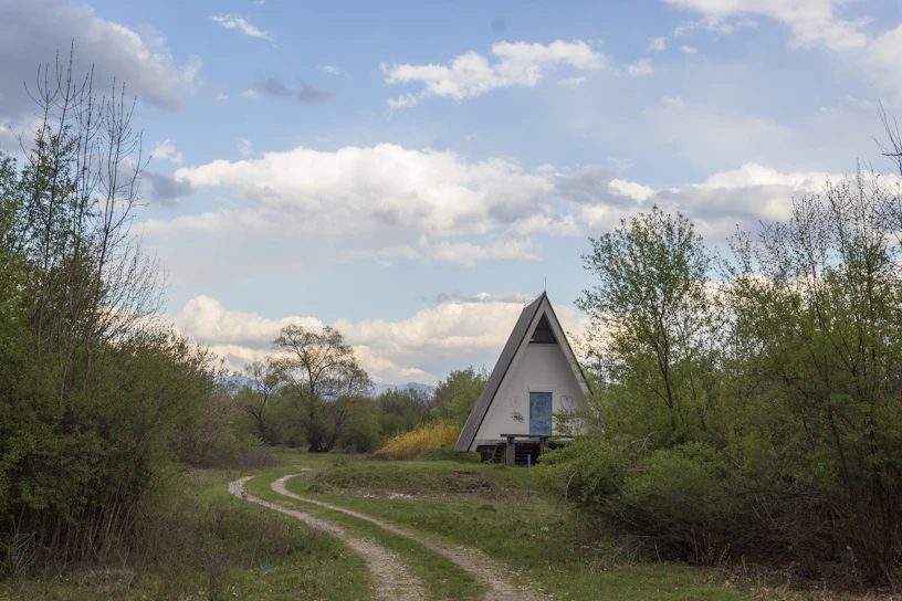 A triangular A-frame house is situated in a natural setting, surrounded by lush green trees and bushes. A narrow dirt path leads to the house, winding through the grassy landscape. Above, the sky is partly cloudy, with patches of blue visible amidst the fluffy white clouds.