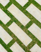 Overlapping rectangular slabs of concrete are placed diagonally, interspersed with green grass in between the gaps. The arrangement forms a visually appealing pattern with contrasting textures.