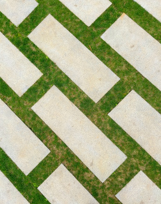 Overlapping rectangular slabs of concrete are placed diagonally, interspersed with green grass in between the gaps. The arrangement forms a visually appealing pattern with contrasting textures.