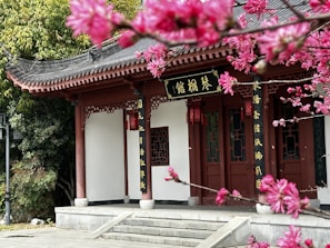 A traditional Asian-style building is surrounded by lush greenery and vibrant pink blossoms. The architectural structure features intricate woodwork and decorative elements on its facade, with a series of steps leading up to the entrance. The combination of natural and man-made beauty creates a serene and harmonious setting.