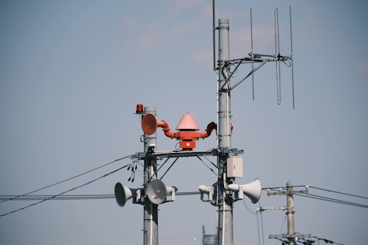 Several utility poles are equipped with various communication devices like loudspeakers, antennas, and a red alert siren. The poles are connected by a network of wires against a clear blue sky.