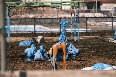 A playground with equipment wrapped in blue tarps, with a fenced boundary in the background. The ground is covered with dry leaves, and there's a visible structure covered with the same material. The scene appears somewhat desolate, suggesting either closure or preparation for protection against weather conditions.