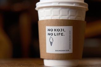 A textured white paper cup with a plastic lid, wrapped in a brown sleeve that features the text 'NO KOJI, NO LIFE.' and a logo of an ice cream cone. The cup is on a wooden surface with a blurred background.