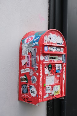 A red mailbox is affixed to a wall and covered with various colorful stickers and graffiti. Some stickers include logos, artistic designs, and text elements, creating an eclectic and urban look.