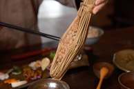 A hand holds a bunch of fermented soybeans wrapped in straw, known as natto. Chopsticks are being used to pick up the soybeans. The background shows a variety of small dishes and a bowl, indicating a meal setting.