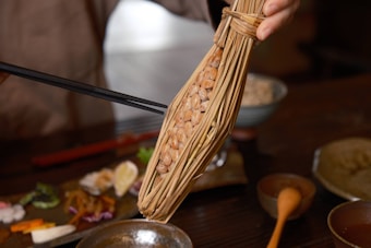 A hand holds a bunch of fermented soybeans wrapped in straw, known as natto. Chopsticks are being used to pick up the soybeans. The background shows a variety of small dishes and a bowl, indicating a meal setting.