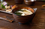 A steaming bowl of miso soup with tofu and seaweed, placed next to chopsticks and a soy sauce dish.