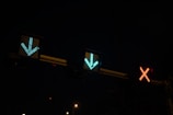 Three illuminated traffic lights are displayed against a dark background. Two green arrows point downward, and a red cross is shown, indicating different directions or statuses.
