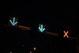 Three illuminated traffic lights are displayed against a dark background. Two green arrows point downward, and a red cross is shown, indicating different directions or statuses.