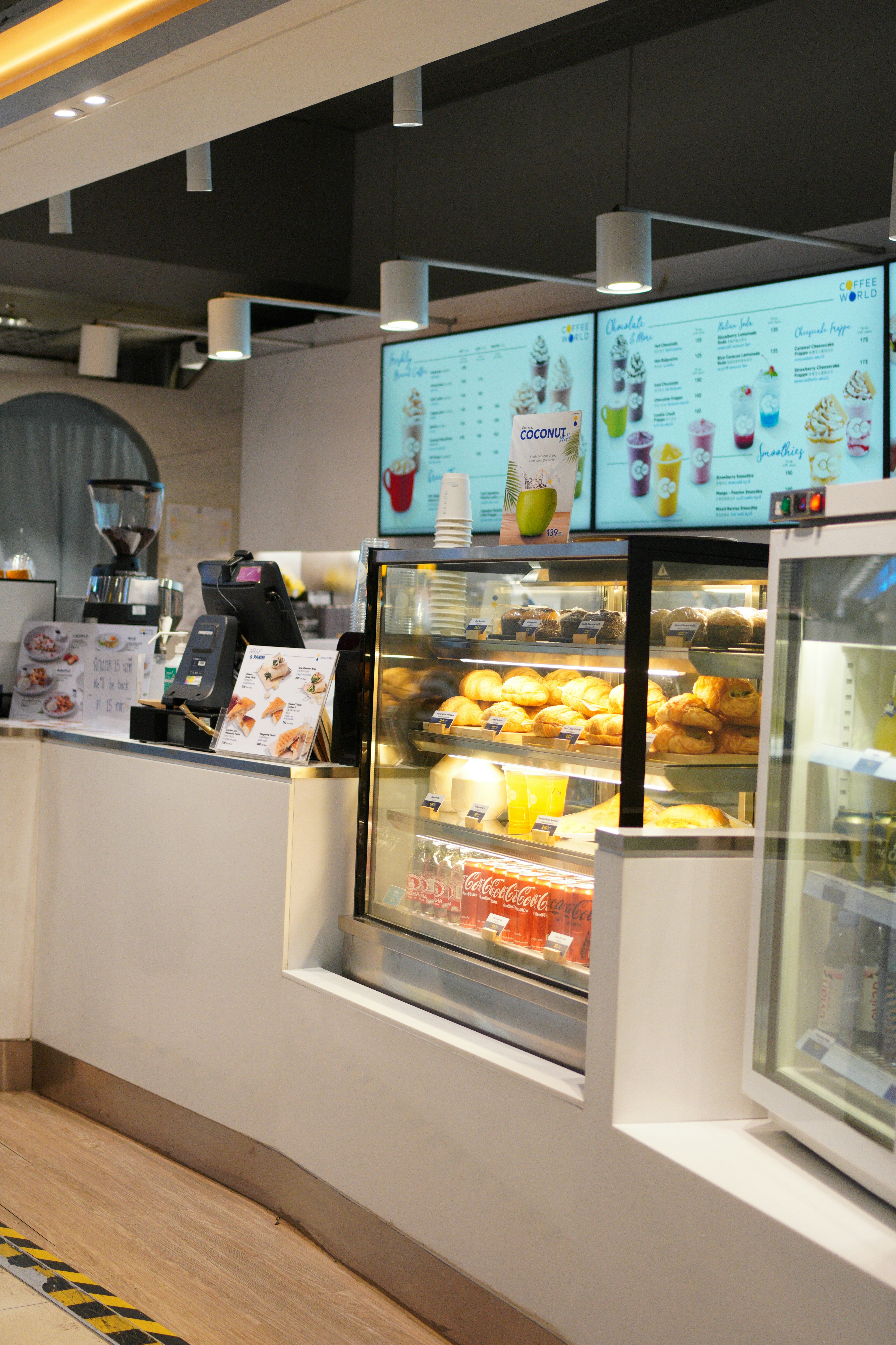 A cafe counter displaying a selection of pastries and beverages with illuminated menu boards above. There is a glass display case housing croissants and bottled drinks, including Coca-Cola. Modern hanging lights provide a soft ambiance.