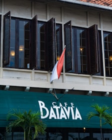 A historic building with wooden shuttered windows stands prominently. The green awning below displays the words 'Cafe Batavia' in white letters. A red and white flag is positioned above the entrance, signifying its location. Two small palm plants are visible on either side of the entrance, adding a touch of greenery.