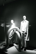 Two mannequins are displayed in a window, both dressed in modern, minimalistic fashion. The scene is accented by large, curved, tube-like structures that add an abstract element to the presentation. The lighting casts dramatic shadows, highlighting the contours of the mannequins and the shapes of the tubes.