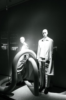 Two mannequins are displayed in a window, both dressed in modern, minimalistic fashion. The scene is accented by large, curved, tube-like structures that add an abstract element to the presentation. The lighting casts dramatic shadows, highlighting the contours of the mannequins and the shapes of the tubes.