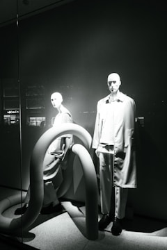 Two mannequins are displayed in a window, both dressed in modern, minimalistic fashion. The scene is accented by large, curved, tube-like structures that add an abstract element to the presentation. The lighting casts dramatic shadows, highlighting the contours of the mannequins and the shapes of the tubes.