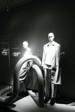 Two mannequins are displayed in a window, both dressed in modern, minimalistic fashion. The scene is accented by large, curved, tube-like structures that add an abstract element to the presentation. The lighting casts dramatic shadows, highlighting the contours of the mannequins and the shapes of the tubes.