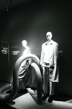 Two mannequins are displayed in a window, both dressed in modern, minimalistic fashion. The scene is accented by large, curved, tube-like structures that add an abstract element to the presentation. The lighting casts dramatic shadows, highlighting the contours of the mannequins and the shapes of the tubes.