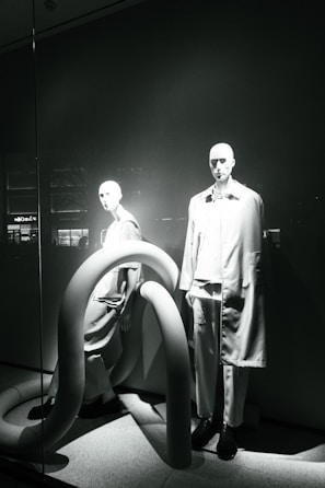 Two mannequins are displayed in a window, both dressed in modern, minimalistic fashion. The scene is accented by large, curved, tube-like structures that add an abstract element to the presentation. The lighting casts dramatic shadows, highlighting the contours of the mannequins and the shapes of the tubes.