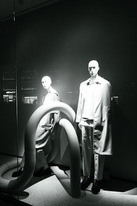 Two mannequins are displayed in a window, both dressed in modern, minimalistic fashion. The scene is accented by large, curved, tube-like structures that add an abstract element to the presentation. The lighting casts dramatic shadows, highlighting the contours of the mannequins and the shapes of the tubes.