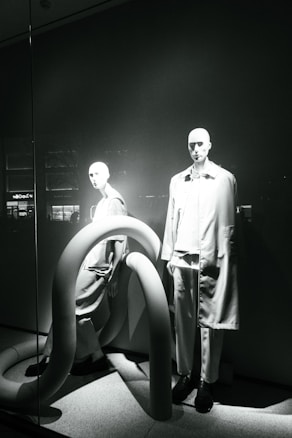 Two mannequins are displayed in a window, both dressed in modern, minimalistic fashion. The scene is accented by large, curved, tube-like structures that add an abstract element to the presentation. The lighting casts dramatic shadows, highlighting the contours of the mannequins and the shapes of the tubes.