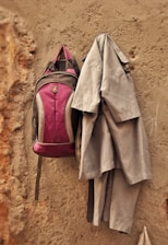 A maroon backpack and a grey uniform hang on a rough, beige wall. The wall has visible textures and signs of wear.