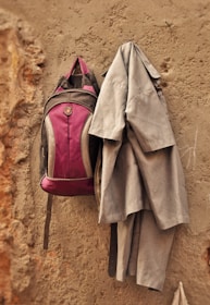 A maroon backpack and a grey uniform hang on a rough, beige wall. The wall has visible textures and signs of wear.