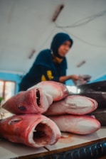 Several large fish heads are laid out on a table, with their mouths open, displaying their sharp teeth. In the background, a person wearing a dark headscarf is blurred, possibly selling fish in a market setting. The room has a bright atmosphere with blue and white tones.