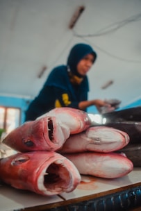 Several large fish heads are laid out on a table, with their mouths open, displaying their sharp teeth. In the background, a person wearing a dark headscarf is blurred, possibly selling fish in a market setting. The room has a bright atmosphere with blue and white tones.