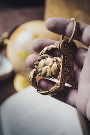 A hand holds a keyring featuring a brass crab-shaped charm and keys. In the background, there is a blurred globe and an open book, suggesting a theme of travel or exploration.