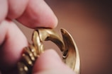 A close-up view of a gold-colored chain link, held between two fingers. The image focuses on the details of the clasp, highlighting its metallic texture and intricate design.