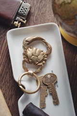 A close-up of a decorative keychain in the shape of a crab, placed on a small white tray. Next to it are two vintage-style keys with inscriptions for good luck. Nearby, a leather-covered lighter and a small section of a globe are visible. The scene is set on a wooden surface with a cigar in the foreground.