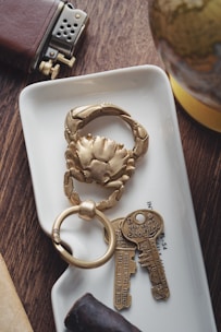 A close-up of a decorative keychain in the shape of a crab, placed on a small white tray. Next to it are two vintage-style keys with inscriptions for good luck. Nearby, a leather-covered lighter and a small section of a globe are visible. The scene is set on a wooden surface with a cigar in the foreground.