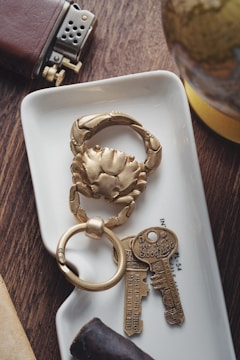 A close-up of a decorative keychain in the shape of a crab, placed on a small white tray. Next to it are two vintage-style keys with inscriptions for good luck. Nearby, a leather-covered lighter and a small section of a globe are visible. The scene is set on a wooden surface with a cigar in the foreground.
