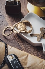 A brass crab-shaped keychain and a pair of keys are placed on a white ceramic dish. Nearby, a black and gold lighter and a car key with black and silver casing are on a wooden table. A yellow section of a globe is slightly visible in the background.
