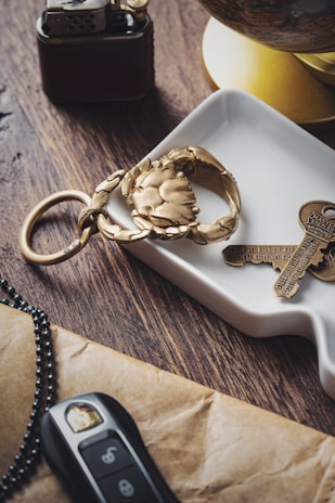 A brass crab-shaped keychain and a pair of keys are placed on a white ceramic dish. Nearby, a black and gold lighter and a car key with black and silver casing are on a wooden table. A yellow section of a globe is slightly visible in the background.