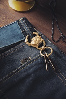 A detailed view of denim jeans with a decorative golden crab keychain attached to the belt loop. The fabric appears dark blue with contrasting orange stitching. A lighter partially peeks out of the front pocket, and a necklace chain is visible in the background on a wooden surface.