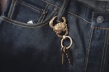 Dark denim jeans featuring a prominent brass keychain shaped like a crab, with a set of keys attached. The fabric's texture and stitching details are visible, highlighting the contrast between the dark denim and the golden keychain.