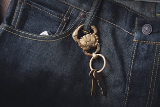 Dark denim jeans featuring a prominent brass keychain shaped like a crab, with a set of keys attached. The fabric's texture and stitching details are visible, highlighting the contrast between the dark denim and the golden keychain.
