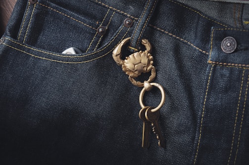 Dark denim jeans featuring a prominent brass keychain shaped like a crab, with a set of keys attached. The fabric's texture and stitching details are visible, highlighting the contrast between the dark denim and the golden keychain.